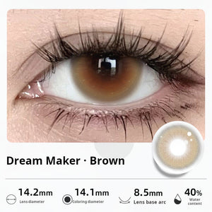 Close-up of an eye wearing "Dream Maker - Brown" contact lenses, featuring a warm hazel-brown center. Specifications: 14.2mm diameter, 14.1mm graphic diameter, 8.5mm base curve, 40% water content.