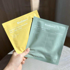 Two Biodance facial mask packets, one yellow and one green, held by a hand against a blurred background.